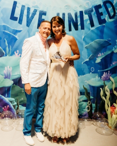 A man and woman in party dress stand in front of an under the sea backdrop. The woman holds an award.