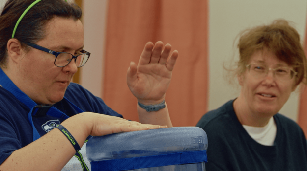 Woman drumming on water jug while another woman watches